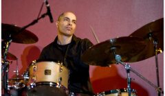 A man is playing a drum set with a neutral expression. The background is a solid maroon color.