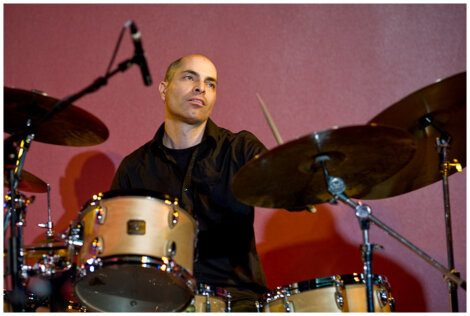 A man is playing a drum set with a neutral expression. The background is a solid maroon color.