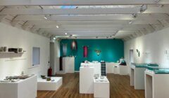 A well-lit gallery room showcases various artworks, including textiles and ceramic pieces, displayed on white pedestals and shelves with a teal accent wall in the background.