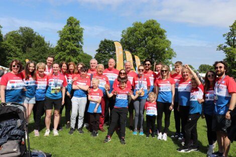 A group of people wearing matching red and blue shirts posing together outdoors on a sunny day, standing on grass with trees and event flags in the background.