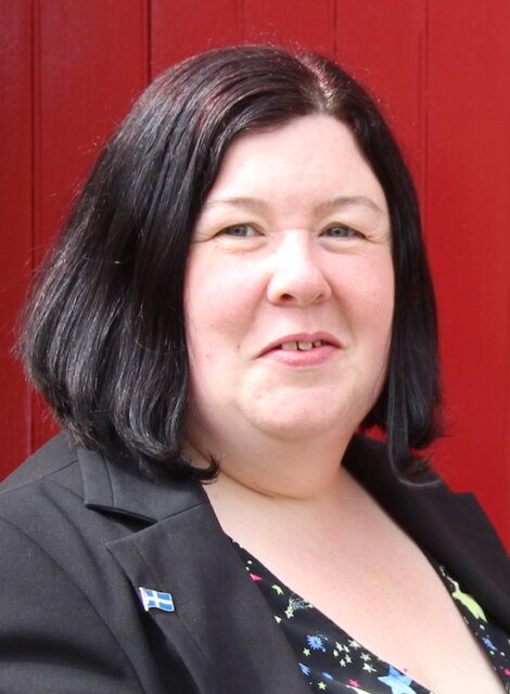 A person with shoulder-length dark hair, wearing a black blazer and a patterned top, stands in front of a red background.