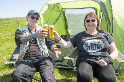 Two people sitting in camping chairs in front of a green tent, smiling and toasting with cans of beer on a sunny day.