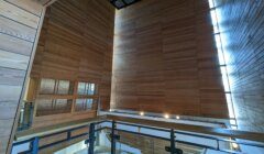 Interior view of a tall, modern wooden building with large wooden panels, a skylight, and a glass railing overlooking a lower level with various wall decorations.