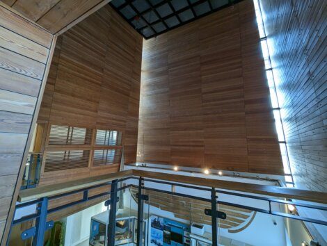 Interior view of a tall, modern wooden building with large wooden panels, a skylight, and a glass railing overlooking a lower level with various wall decorations.