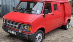 A red Bedford van with a distinctive boxed shape and front grille is parked on a concrete surface near a corrugated metal building. The license plate reads "C834 AAS.