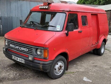 A red Bedford van with a distinctive boxed shape and front grille is parked on a concrete surface near a corrugated metal building. The license plate reads "C834 AAS.