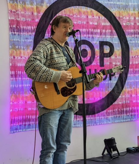 A man in a patterned sweater plays an acoustic guitar and sings into a microphone on stage, standing before a colorful backdrop with text partially displaying "POP UP.