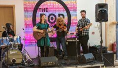 A band performs on stage with a drummer, guitarist, violinist, and bassist in front of a backdrop that reads "The Pop.