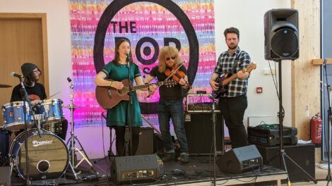 A band performs on stage with a drummer, guitarist, violinist, and bassist in front of a backdrop that reads "The Pop.