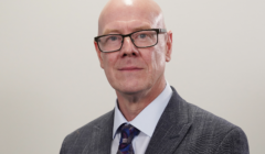 Bald man with glasses wearing a gray suit, light blue shirt, and patterned tie, standing against a plain background.