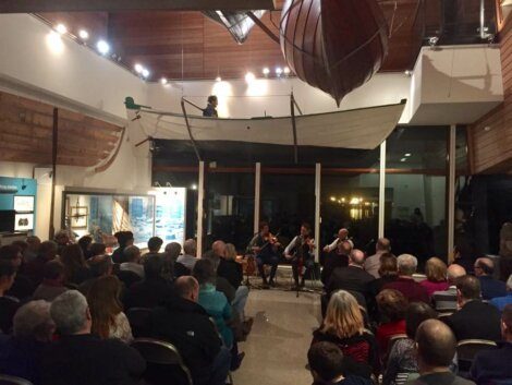 A group of people seated in a room with a boat suspended above. Four individuals are engaged in conversation or performance at the front, with an audience watching attentively.