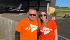 Two people wearing orange MS Society shirts stand in front of an airplane hangar, smiling at the camera.