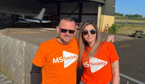 Two people wearing orange MS Society shirts stand in front of an airplane hangar, smiling at the camera.