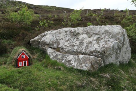 A large, weathered rock and a small red house-like structure with a grass roof are situated on a grassy hillside surrounded by sparse vegetation.