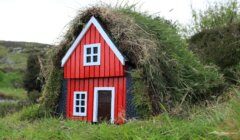 A small red house with a white trim and roof covered in grass sits amidst greenery.