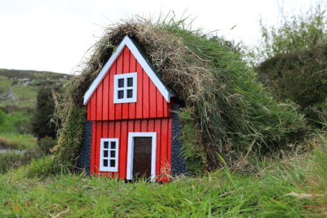 A small red house with a white trim and roof covered in grass sits amidst greenery.