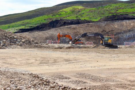 A construction site featuring two excavators, one orange and one black, operating on dirt and rocks near hills with green vegetation.