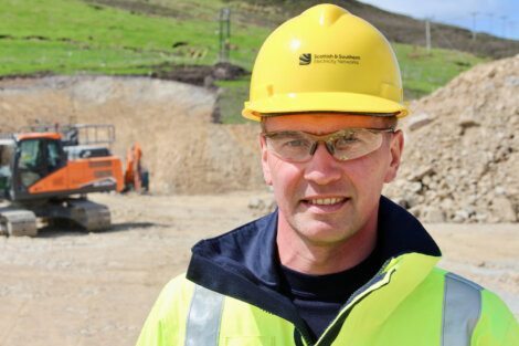 A person wearing a yellow hard hat and high-visibility jacket stands at a construction site with an excavator in the background.