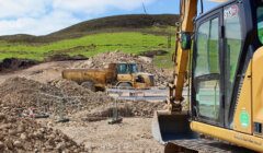 Construction site with heavy equipment, including a bulldozer and a dump truck, working on a rocky terrain. A grassy hill and a wind turbine can be seen in the background.