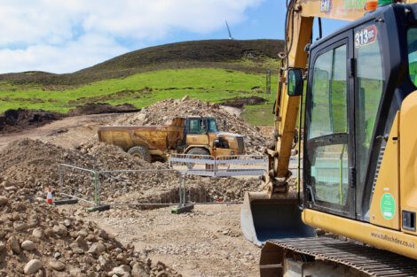 Construction site with heavy equipment, including a bulldozer and a dump truck, working on a rocky terrain. A grassy hill and a wind turbine can be seen in the background.