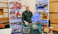 A man stands behind a display table with fishing equipment, promotional materials, and a sign reading "Certified Sustainable Seafood MSC," with posters about sustainable shellfish and local fishermen in the background.