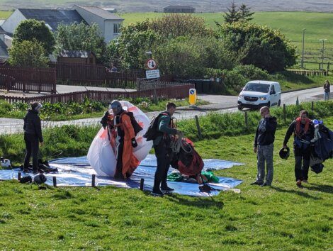 Parachutists pack their gear on a grassy area near a road, with a white van and houses in the background.