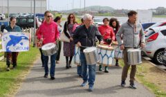 A group of people walking outdoors, some playing drums. Others hold signs, one reads "Leave It in the Ground." Several parked cars and buildings are in the background.