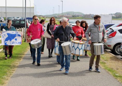 A group of people walking outdoors, some playing drums. Others hold signs, one reads "Leave It in the Ground." Several parked cars and buildings are in the background.