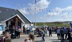 A crowd gathers outside a building with red trim and a flagpole. Bunting decorates the scene. The sky is partly cloudy, and hills are visible in the background.