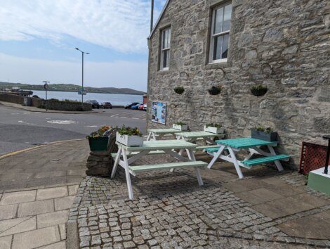 Outdoor seating area with two picnic tables next to a stone building, overlooking a parking lot and a view of the sea in the distance.