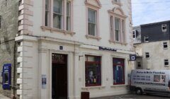 A white building with large windows displays a sign reading "Royal Bank of Scotland." A parked van advertising painting and decorating services is positioned to the right of the building’s entrance.