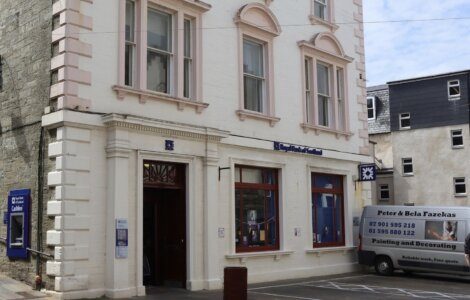 A white building with large windows displays a sign reading "Royal Bank of Scotland." A parked van advertising painting and decorating services is positioned to the right of the building’s entrance.