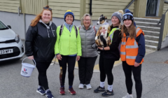 Five women, dressed in casual outdoor attire, stand together smiling for a photo. One woman is holding a dog. Another holds a collection bucket. They are outdoors near a building with steps.