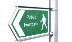 Green and white sign on a post indicating a public footpath with a pedestrian icon pointing left.