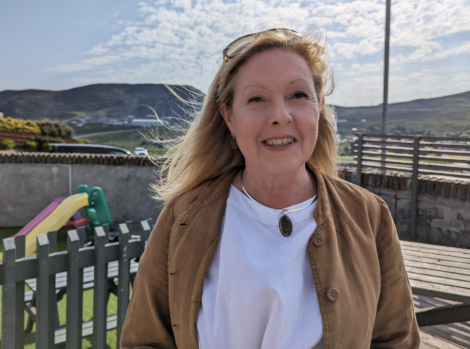 Woman standing outdoors smiling with mountains in the background and children's playground equipment visible behind a fence.