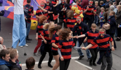 A group of people in matching red and black striped shirts dance in the street during a crowded parade. A person on stilts is visible in the background. Spectators line both sides of the street.