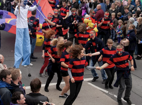 A group of people in matching red and black striped shirts dance in the street during a crowded parade. A person on stilts is visible in the background. Spectators line both sides of the street.