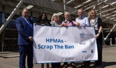 A group of six people stand outdoors holding a banner that reads, "HPMAs - Scrap The Ban." They are in front of a building with modern architectural features.