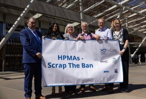 A group of six people stand outdoors holding a banner that reads, "HPMAs - Scrap The Ban." They are in front of a building with modern architectural features.