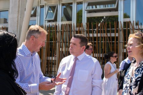 Four people are outside a building having a conversation. One man is speaking while gesturing with his hands. The others are listening attentively. There are additional people in the background.