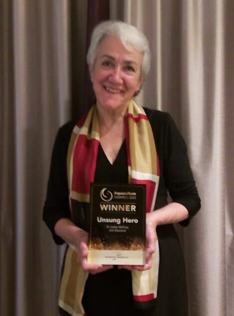 A person with white hair smiles while holding a plaque titled "WINNER Unsung Hero" at the Aquaculture Awards 2020.