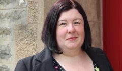 A woman with shoulder-length dark hair stands in front of a stone wall, wearing a black blazer with a small flag pin and a patterned top.