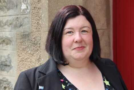 A woman with shoulder-length dark hair stands in front of a stone wall, wearing a black blazer with a small flag pin and a patterned top.