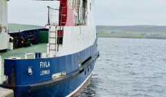 A ferry named "Fivla Lerwick" is docked at a harbor with land visible in the background. The ferry's hull is blue and white with red accents.