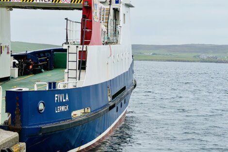 A ferry named "Fivla Lerwick" is docked at a harbor with land visible in the background. The ferry's hull is blue and white with red accents.