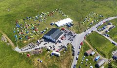 Aerial view of a rural event site with numerous small tents scattered around several large tents and a building, surrounded by fields and a few parked vehicles along adjacent roads.