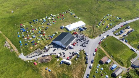 Aerial view of a rural event site with numerous small tents scattered around several large tents and a building, surrounded by fields and a few parked vehicles along adjacent roads.