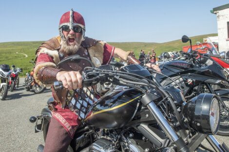 A person wearing a Viking costume sits on a motorcycle, shouting with an open mouth. Multiple motorcycles and people are visible in the background on a sunny day.