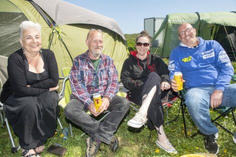 Four people are sitting outside tents at a campsite, smiling and holding drinks.