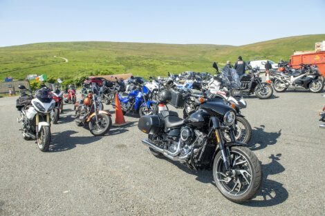 A group of motorcycles parked on a paved area with green hills in the background under a clear sky.